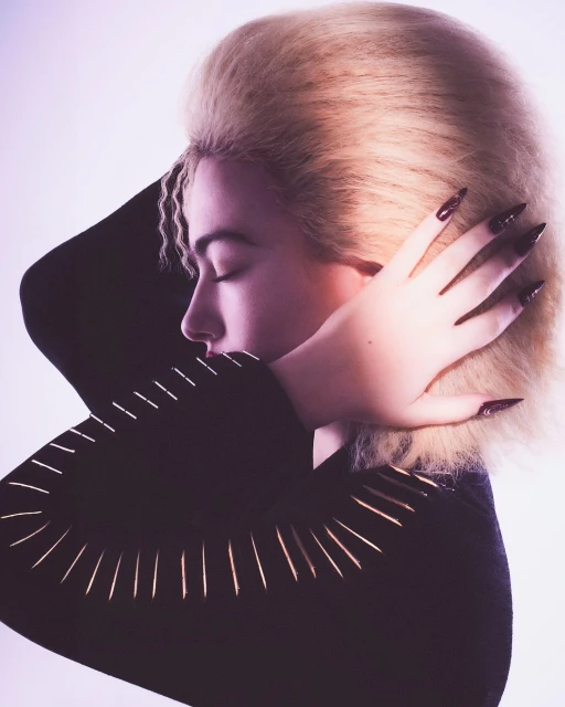 Close-up portrait of a woman with blonde bouffant hair, long dark nails, eyes closed, wearing a black top with gold accents, against a light background.
