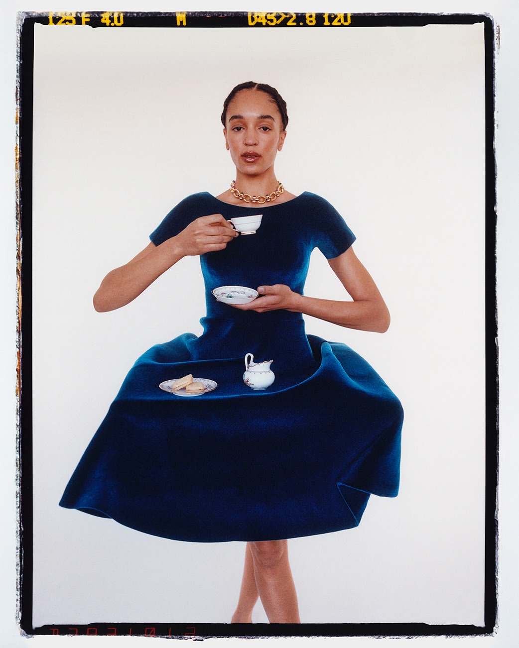 Woman in large blue dress holds tea cup and saucer against white backdrop in portrait shot with visible film border.