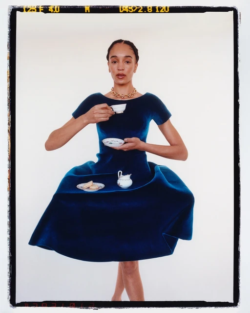 Woman in large blue dress holds tea cup and saucer against white backdrop in portrait shot with visible film border.