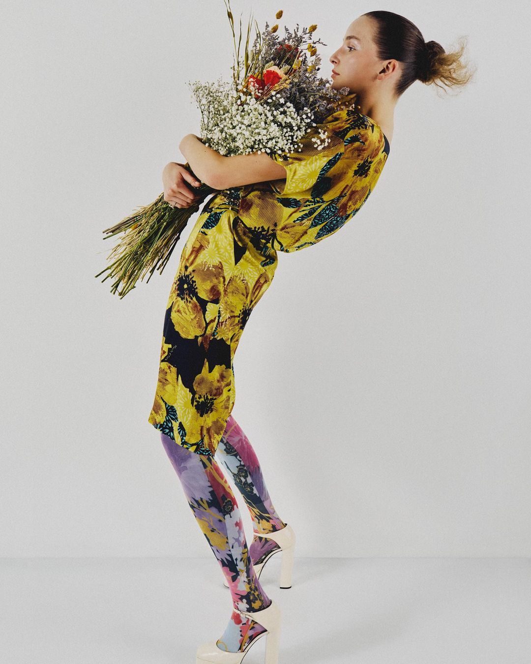 A slender woman poses indoors, wearing a yellow floral dress and floral leggings while holding a bouquet of flowers against a white backdrop.