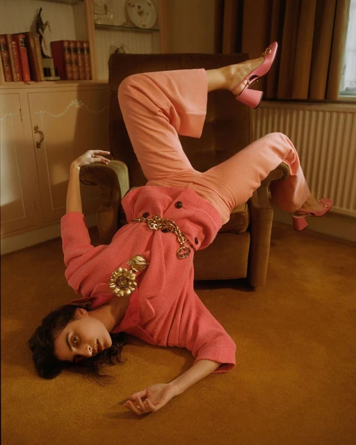 Woman in pink suit, pants, and platform heels strikes a creative pose indoors on an ochre rug with neutral backdrop and soft lighting.