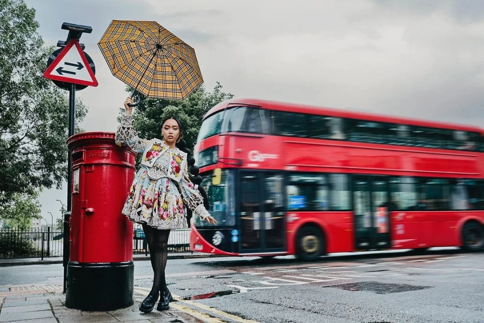 A woman stands on a street corner holding an umbrella next to a red postbox with a red double-decker bus passing by.