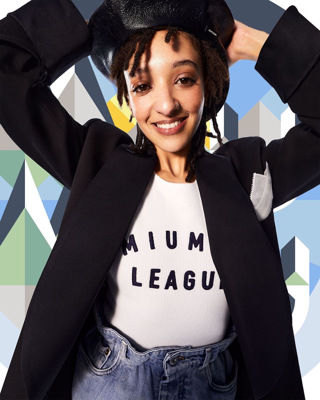 Smiling woman in a black blazer and beret poses against a geometric backdrop, exuding casual style and confidence in a studio setting.