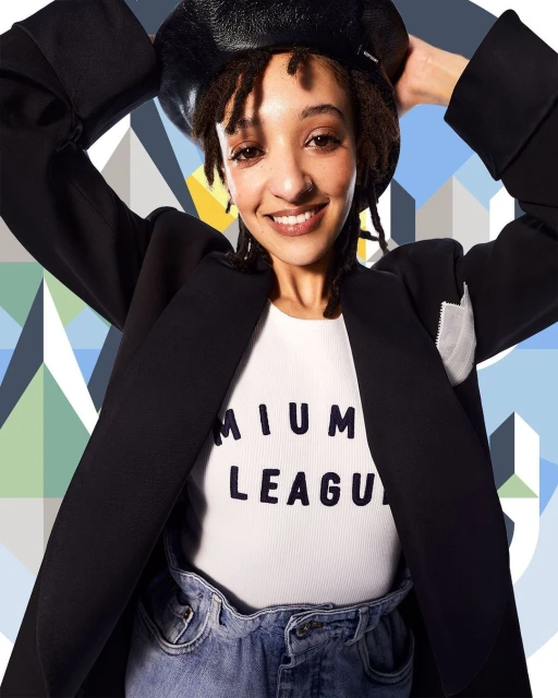 Smiling woman in a black blazer and beret poses against a geometric backdrop, exuding casual style and confidence in a studio setting.
