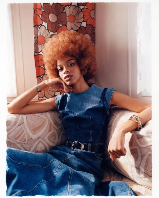A woman with an afro hairstyle wearing a denim jumpsuit and jewelry poses indoors in front of a floral patterned wall hanging.