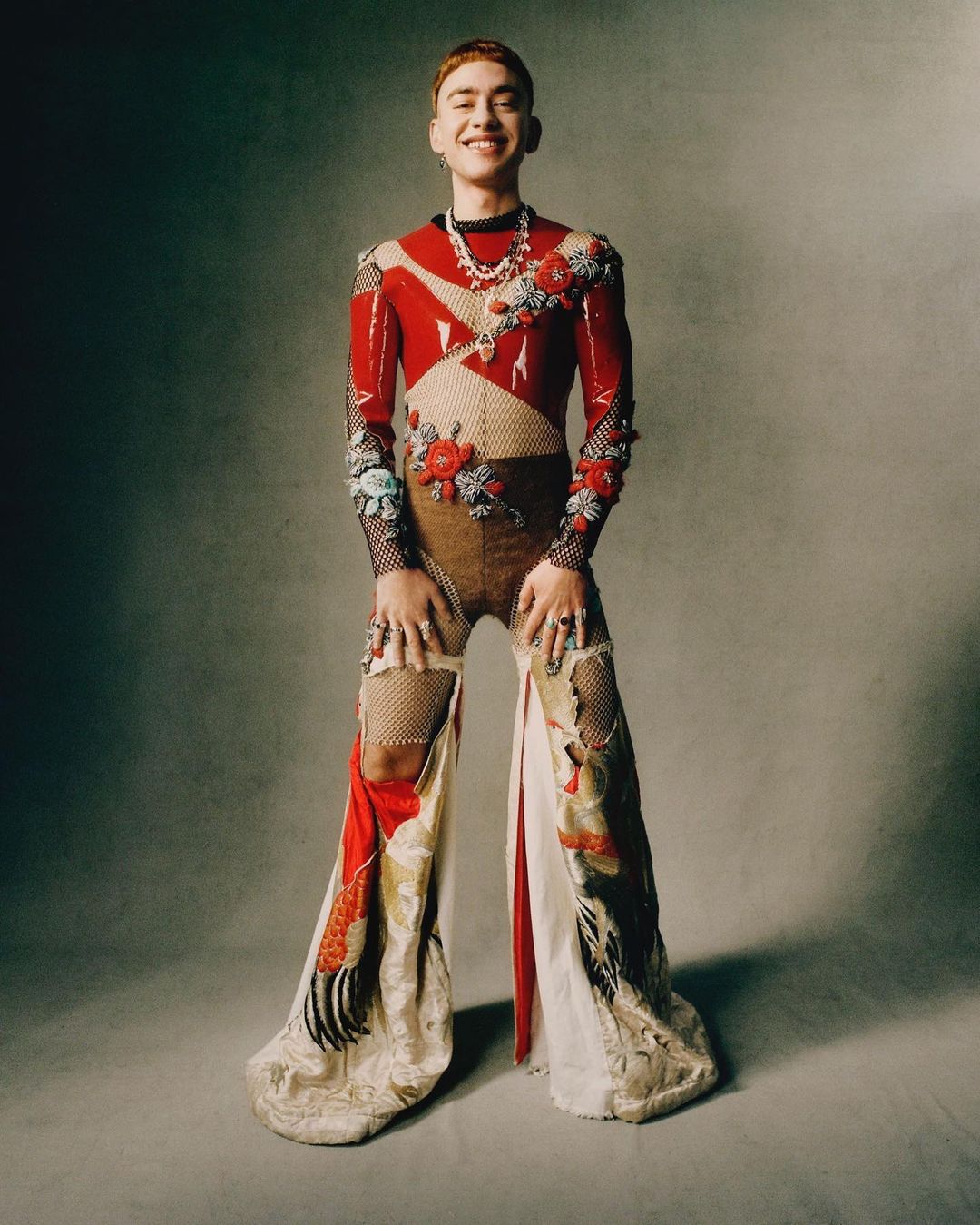 Full-length portrait of a young adult with red hair wearing a decorated, patterned outfit with jewelry, posing in a studio.