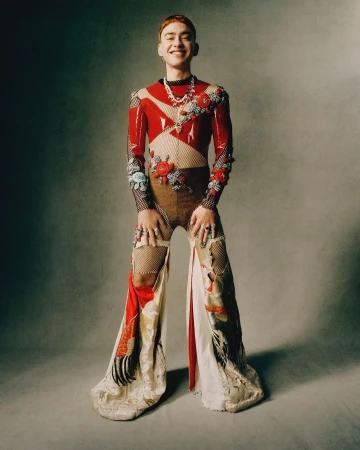 Full-length portrait of a young adult with red hair wearing a decorated, patterned outfit with jewelry, posing in a studio.