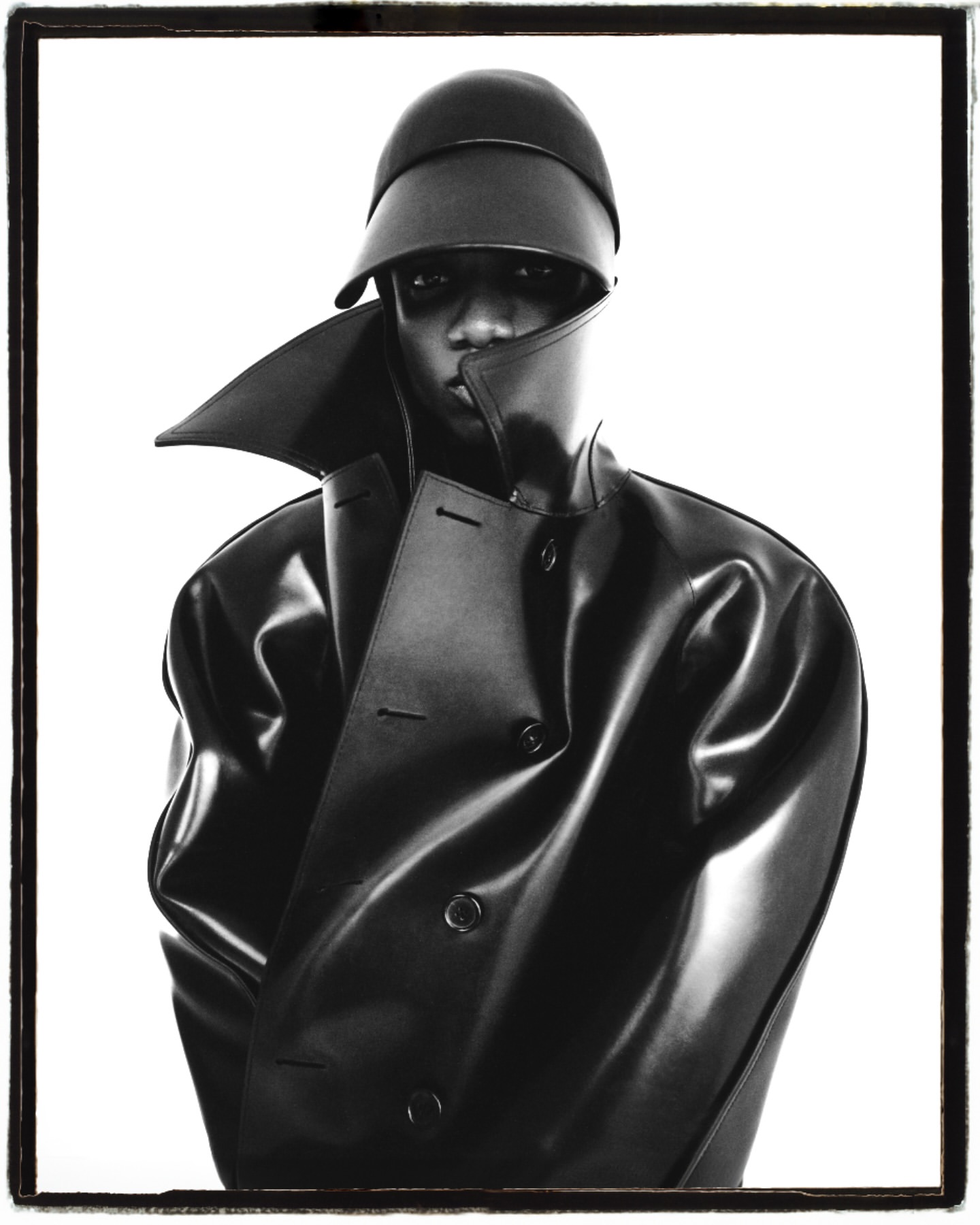 Striking monochrome studio portrait of a Black adult male wearing a black hat and a shiny black trench coat against a stark white backdrop.