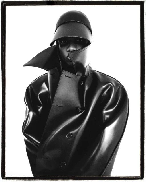 Striking monochrome studio portrait of a Black adult male wearing a black hat and a shiny black trench coat against a stark white backdrop.
