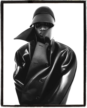 Striking monochrome studio portrait of a Black adult male wearing a black hat and a shiny black trench coat against a stark white backdrop.