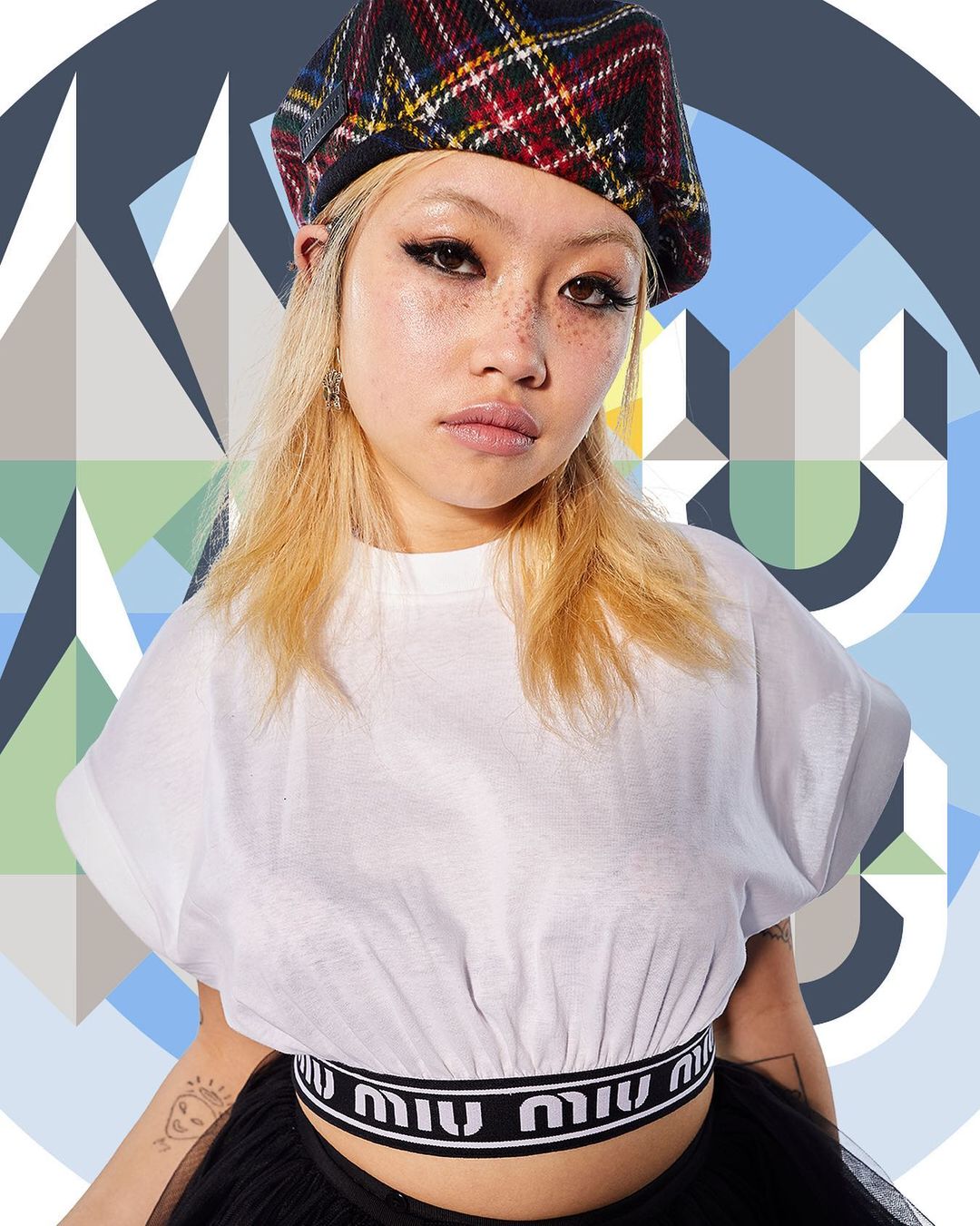 A young woman with blonde hair and a plaid beret poses in front of a geometric blue background, wearing a white top with logo waistband.