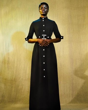 Full-length portrait of a dark-skinned female model in a long-sleeved, ankle-length black dress with a high collar and white button details.