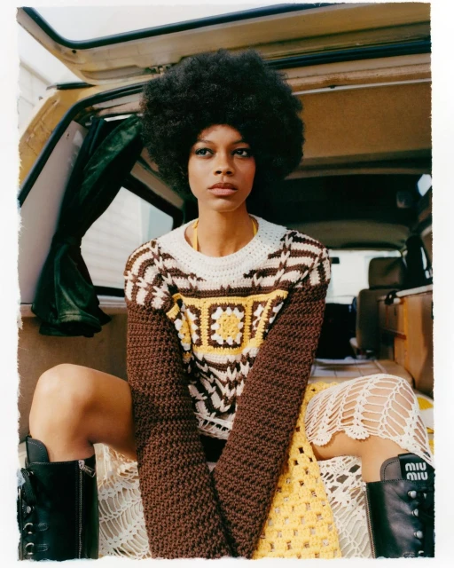 A fashionable young woman with an afro hairstyle poses inside the back of a van, wearing a crochet sweater and skirt, and black boots.
