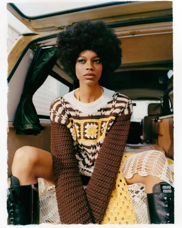 A fashionable young woman with an afro hairstyle poses inside the back of a van, wearing a crochet sweater and skirt, and black boots.
