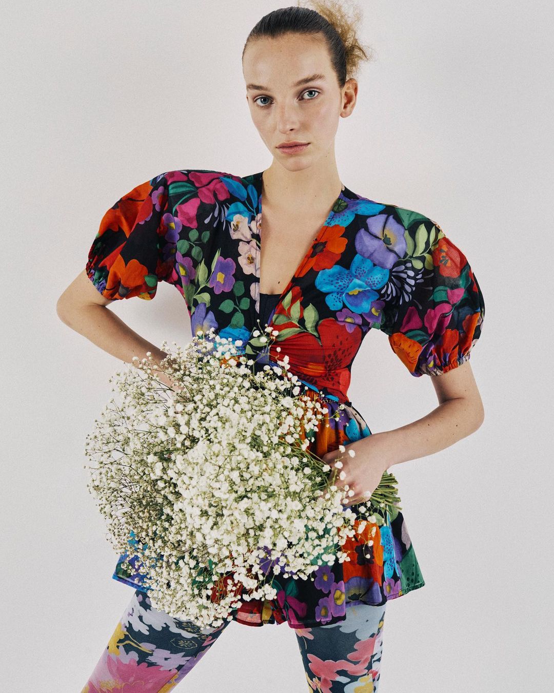 A woman in colorful floral print top and leggings holds a bouquet of baby's breath flowers against a white background.