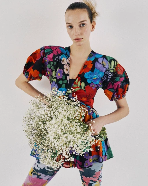 A woman in colorful floral print top and leggings holds a bouquet of baby's breath flowers against a white background.