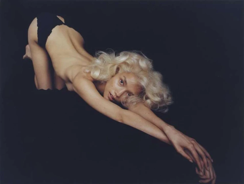 A woman with blonde hair is in a crawling position on a dark fabric in a studio, wearing a black swimsuit bottom. Her arms are extended.