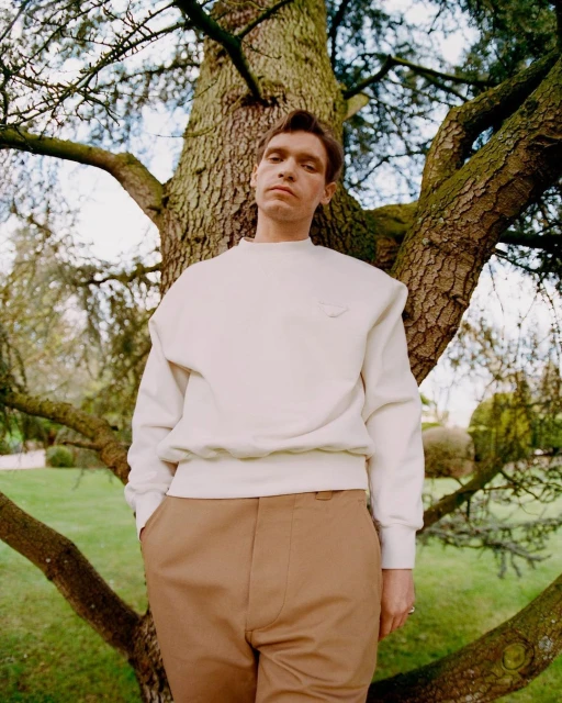 A young adult man stands near a large tree in a natural outdoor setting wearing a white sweatshirt and tan pants in daylight.