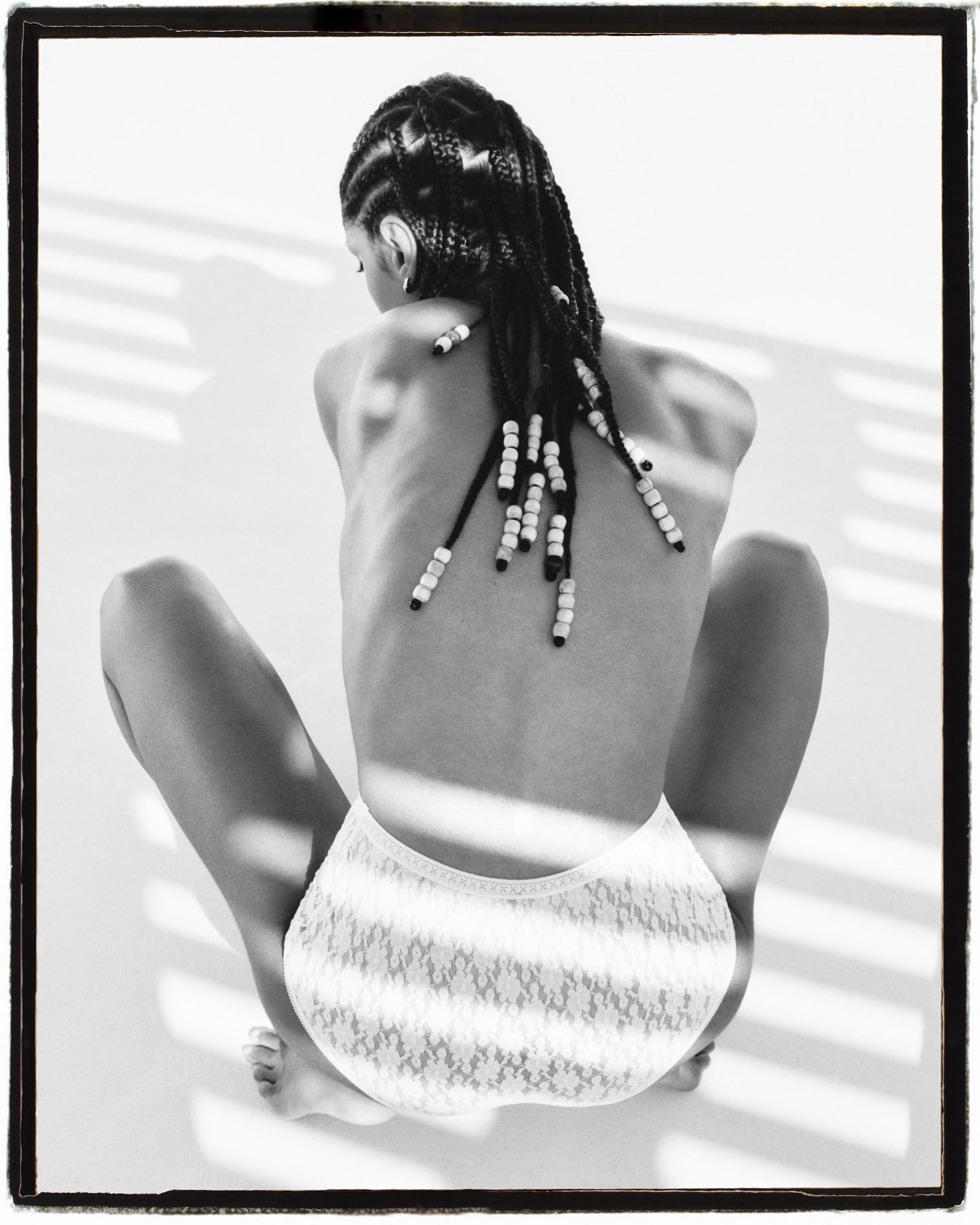 Back view of a young adult woman with beaded braids wearing white lace briefs sitting on a white surface with shadow pattern.