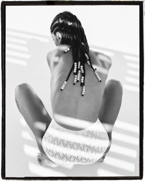 Back view of a young adult woman with beaded braids wearing white lace briefs sitting on a white surface with shadow pattern.