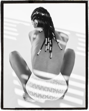 Back view of a young adult woman with beaded braids wearing white lace briefs sitting on a white surface with shadow pattern.