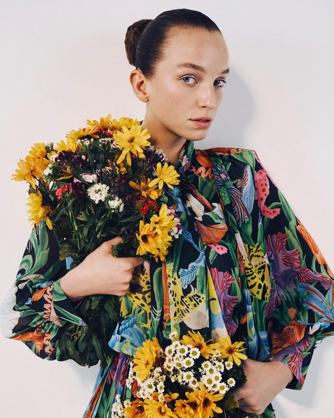 A woman in a floral print top holding bouquets of yellow flowers against a plain white background by Nick Thompson.
