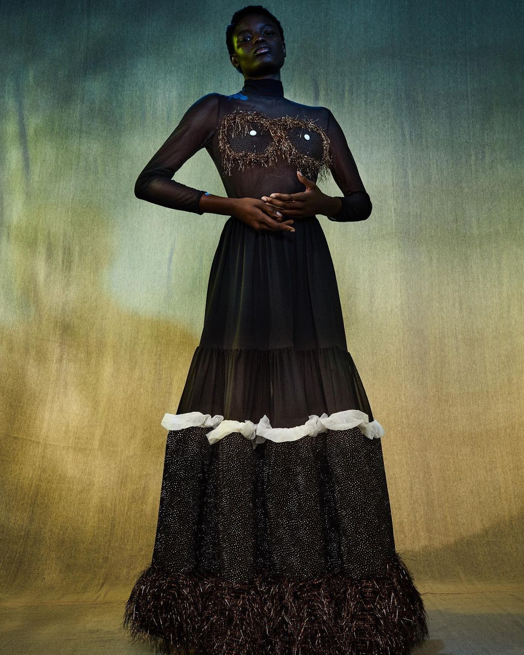 Full-length studio shot: Woman in black sheer gown with ornate bodice, hands clasped at waist, against a mottled green and gold backdrop.