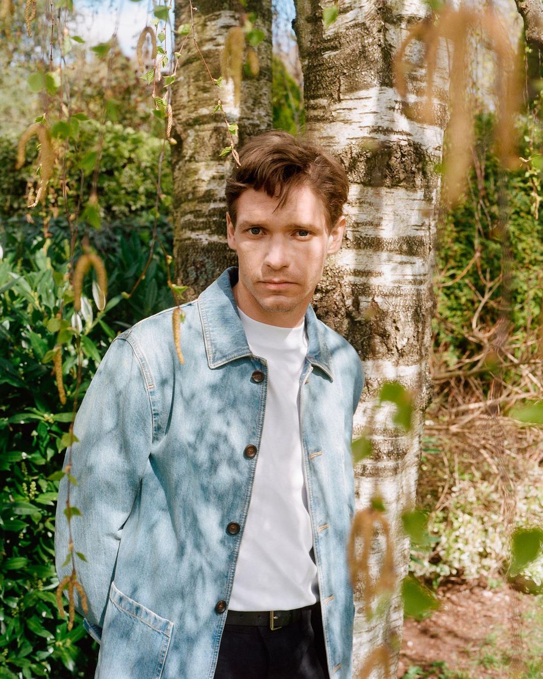 Portrait of a young adult man wearing a denim jacket and white t-shirt standing by birch trees in a lush green outdoor setting.