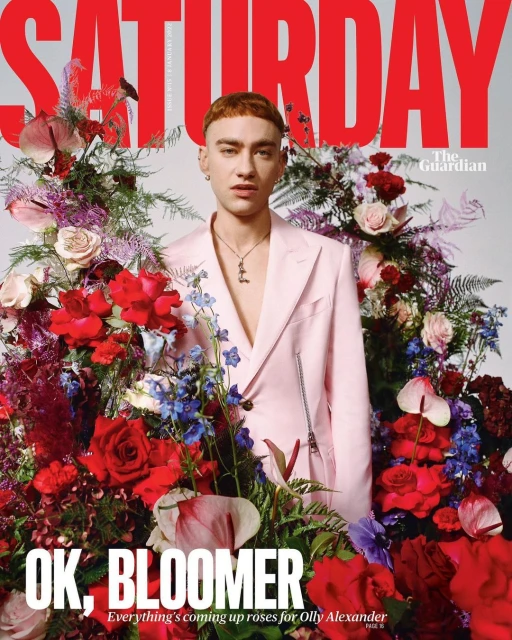 Magazine cover featuring Olly Alexander in a pink suit surrounded by lush red, pink, and purple flowers, with 'Saturday' in bold red text.