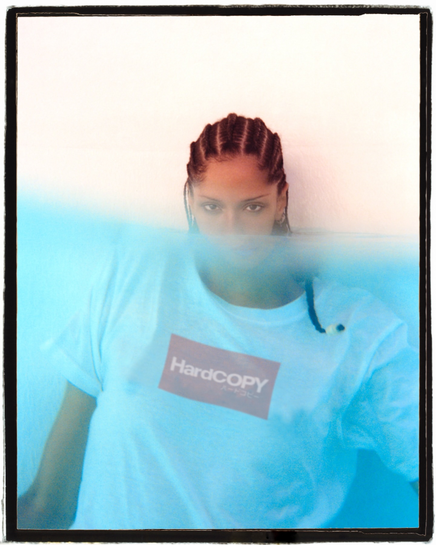 Portrait of a woman with braids wearing a Hardcopy branded t-shirt, partially submerged in blue water against a white background.