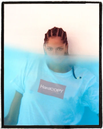 Portrait of a woman with braids wearing a Hardcopy branded t-shirt, partially submerged in blue water against a white background.
