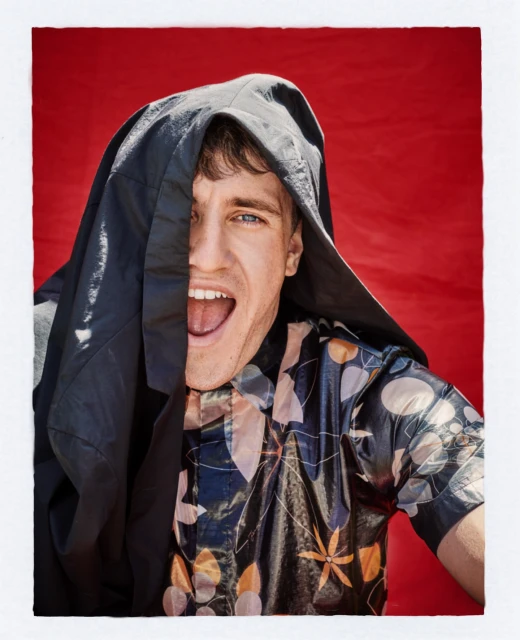 Portrait of a young adult man with an open mouth, partially covered by a black hooded garment, set against a solid red background.