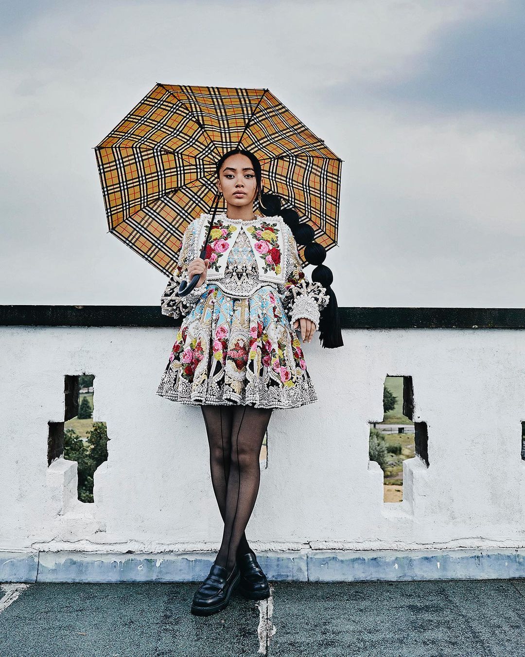 Woman in a floral dress and black tights holding a checkered umbrella on a rooftop with a cloudy sky in the background.