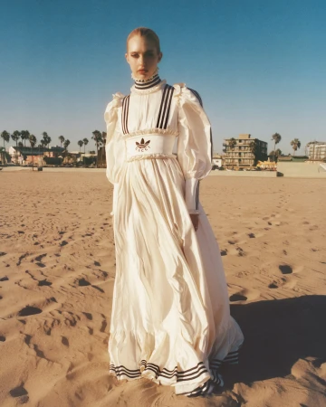Model in a long white ruffled dress standing on a sandy beach under a clear sky by Nick Thompson