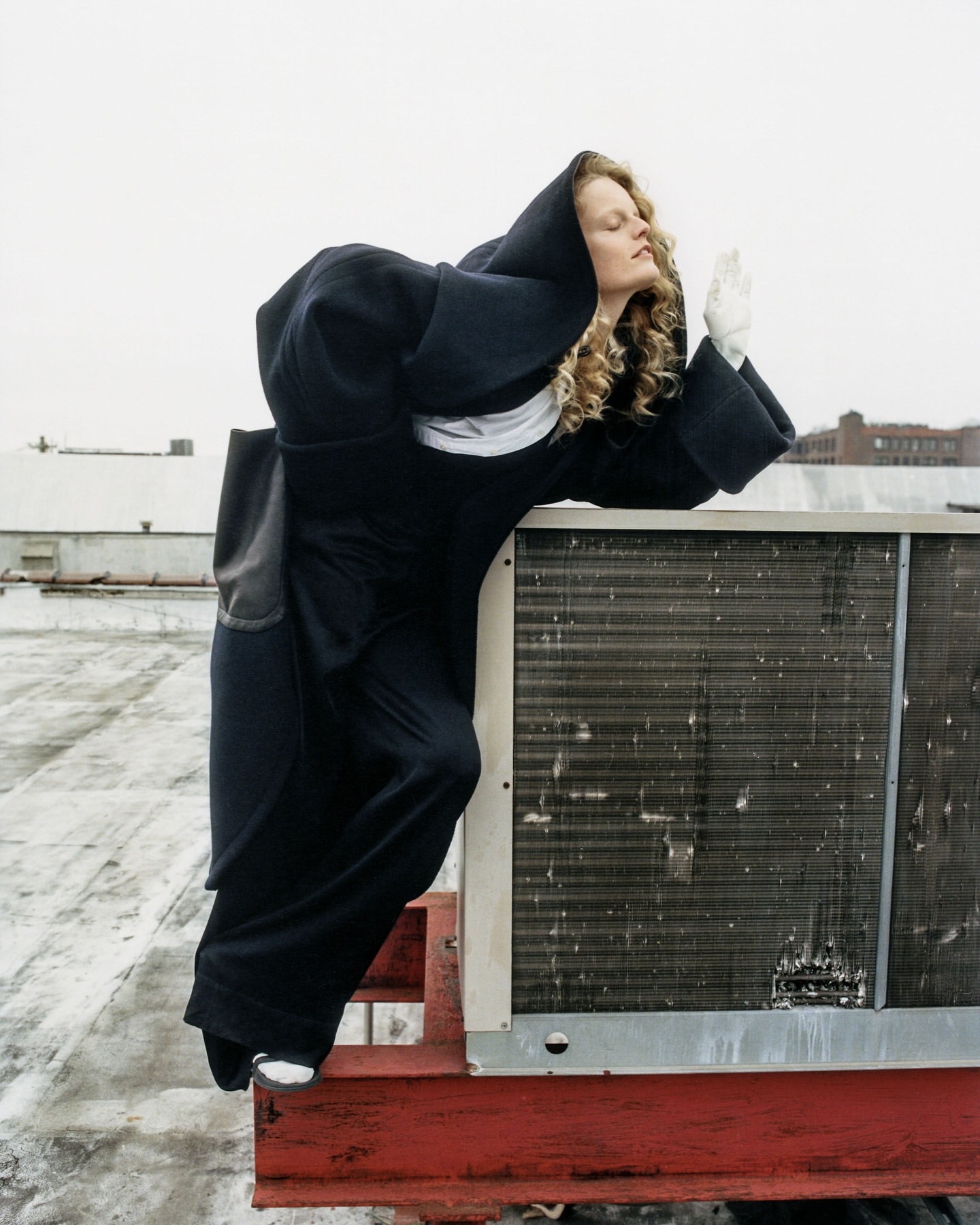 Adult female model in a dark hooded garment poses on top of a building with an HVAC system against a blurred urban background.