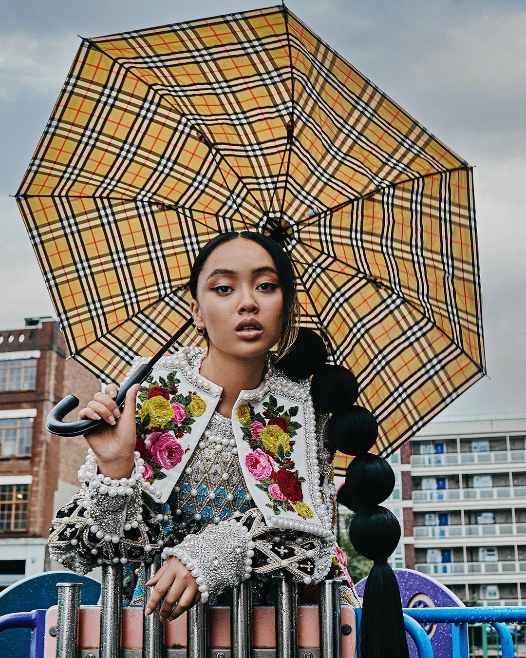 Portrait of an Asian woman under a Burberry umbrella, wearing an embellished jacket, outdoors on a playground, set against building backdrop.