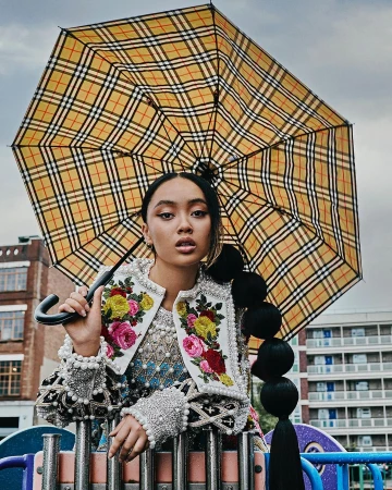 Portrait of an Asian woman under a Burberry umbrella, wearing an embellished jacket, outdoors on a playground, set against building backdrop.