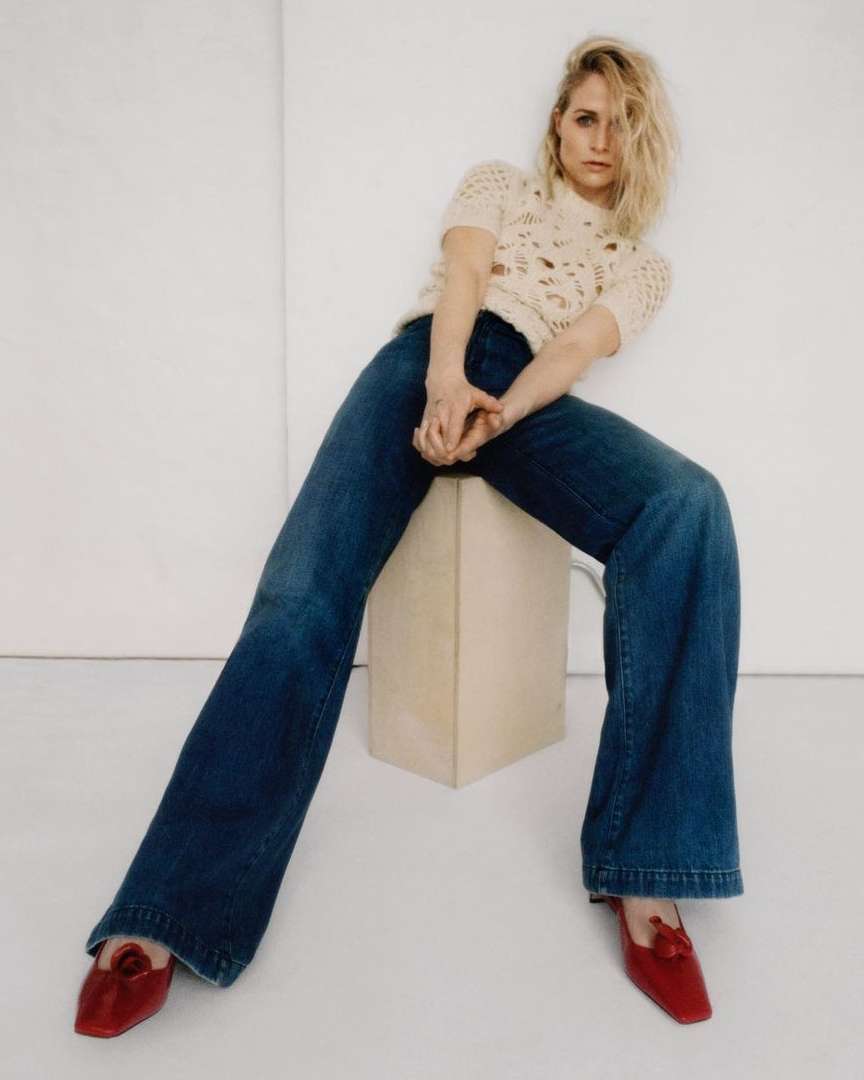 Blonde woman in a cream knit top and flared jeans posing in a minimalist studio, seated on a cream cube with red flats.