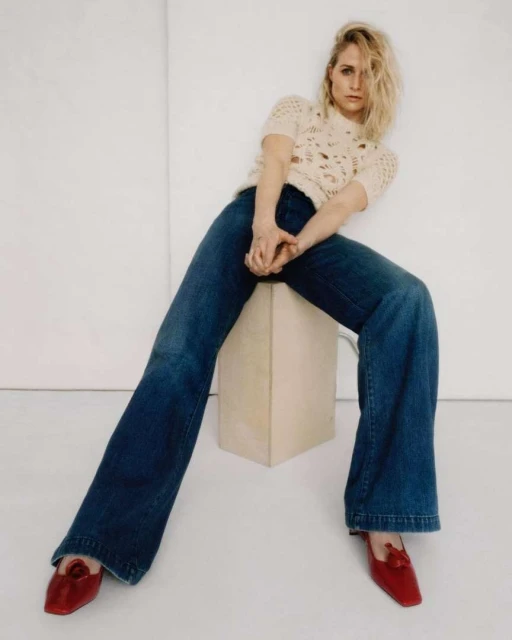 Blonde woman in a cream knit top and flared jeans posing in a minimalist studio, seated on a cream cube with red flats.