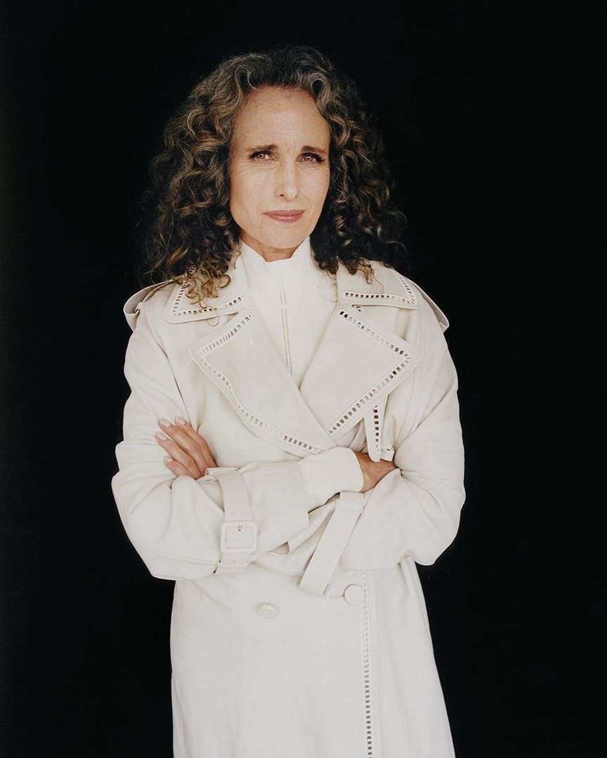 A portrait of an adult woman with curly hair, wearing a light-colored coat, posing with arms crossed against a black background.