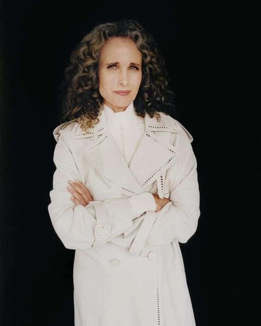A portrait of an adult woman with curly hair, wearing a light-colored coat, posing with arms crossed against a black background.