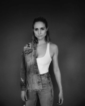 Portrait of a woman wearing a white tank top and denim jeans with a denim jacket draped over one shoulder against a dark background.