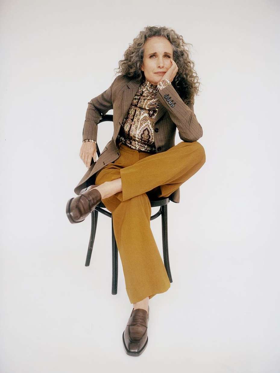 Mature woman with curly gray hair sits cross-legged on a black chair in a photo studio wearing a brown blazer and golden pants.