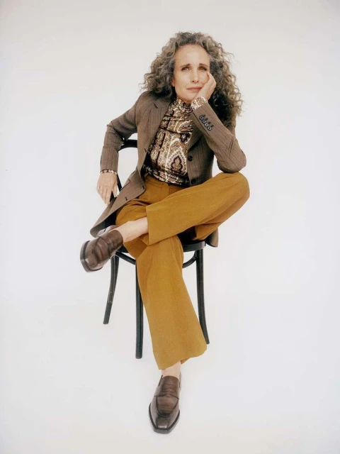 Mature woman with curly gray hair sits cross-legged on a black chair in a photo studio wearing a brown blazer and golden pants.