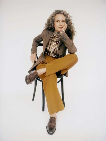Mature woman with curly gray hair sits cross-legged on a black chair in a photo studio wearing a brown blazer and golden pants.