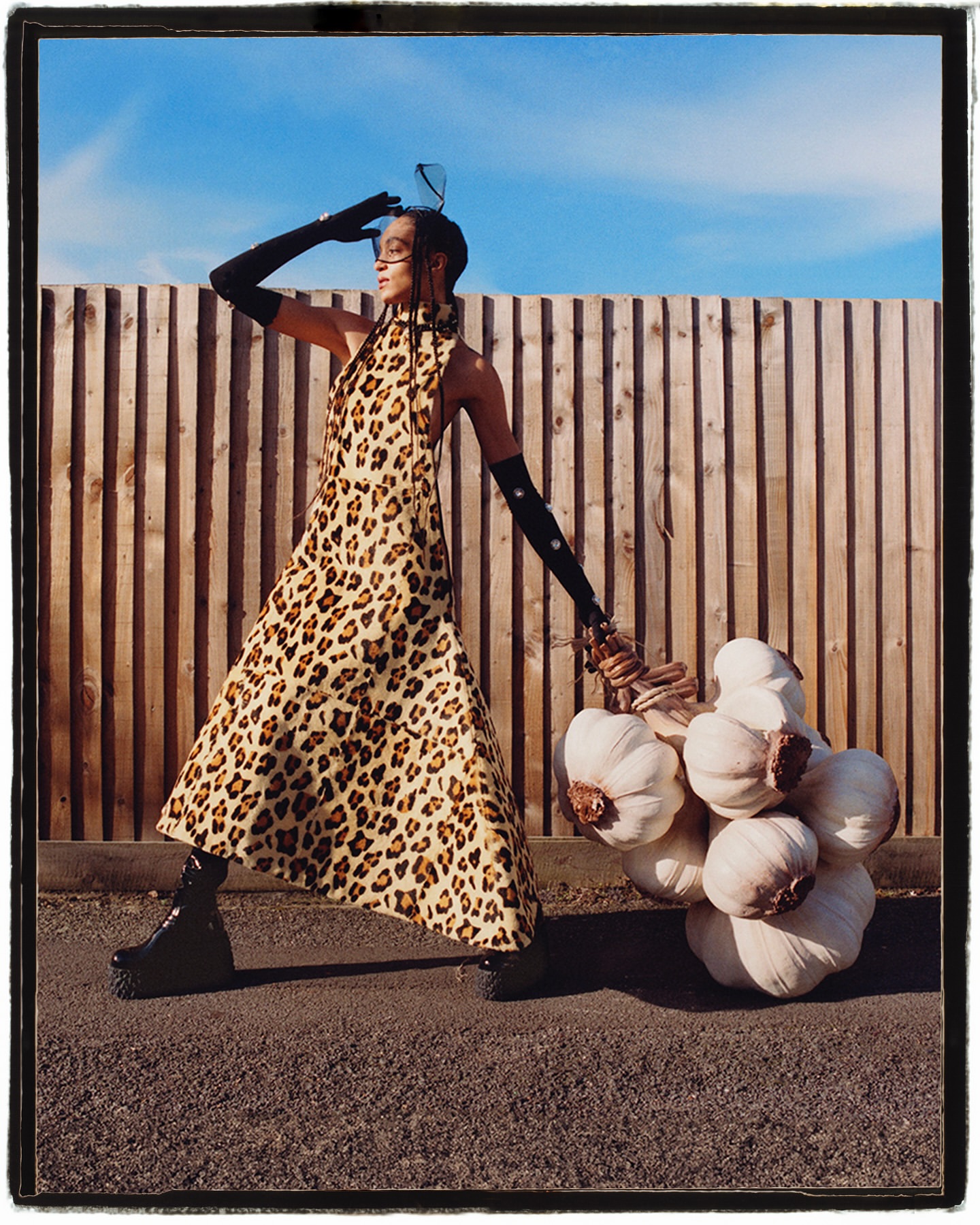 A woman in a leopard-print dress and platform boots poses outdoors, carrying a bunch of garlic in front of a wooden fence.