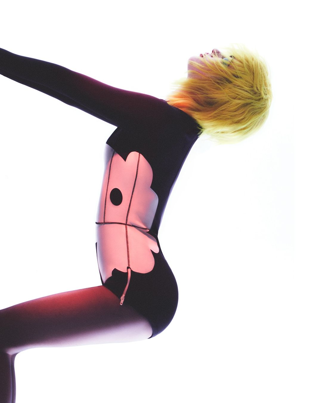 Woman with yellow hair in a black and pink patterned bodysuit against a white backdrop, angled to the left, arms raised, studio setting.
