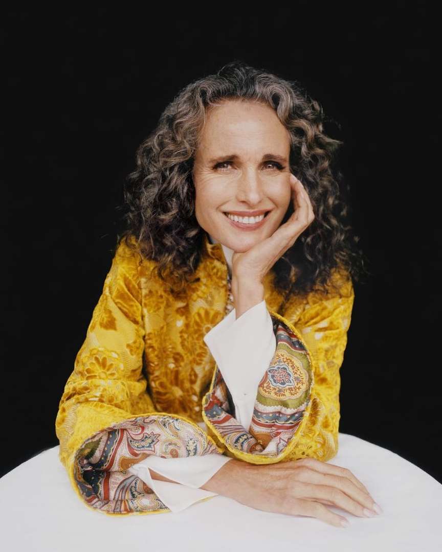 Portrait of Andie MacDowell in a patterned yellow jacket with curly gray hair, smiling with her chin resting on her hand.