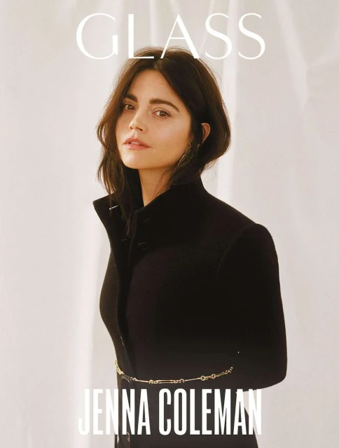 Jenna Coleman poses in a black top and gold chain belt against a neutral background for a magazine cover. Portrait shot.