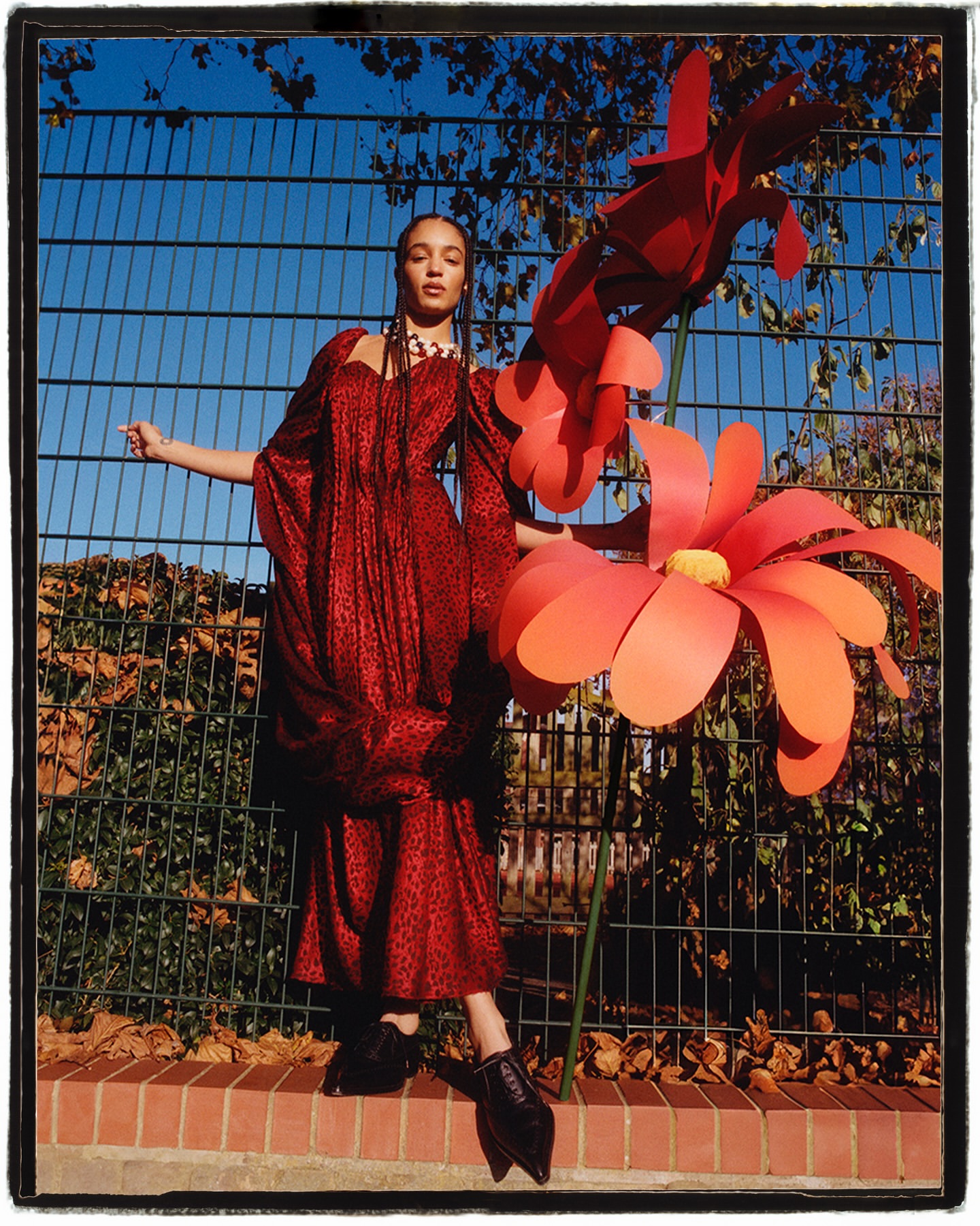 A woman in a patterned red dress stands outdoors next to large paper flower sculptures against a fence and blue sky in the background.